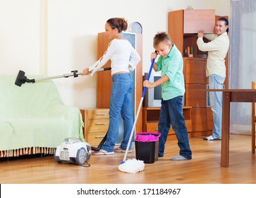 Ordinary Family Of Three With Teenager Doing Housework With  Cleaning Equipment In Living Room