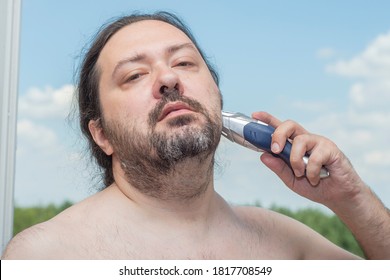 An ordinary middle-aged adult man with ponytail hair shaves his beard with an electric trimmer. Man on a light background.