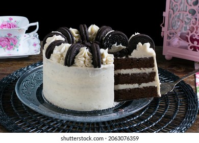Oreo cookie cake with cream cheese frosting on plate on black tablecloth, black background, cup of flowers and pink lantern