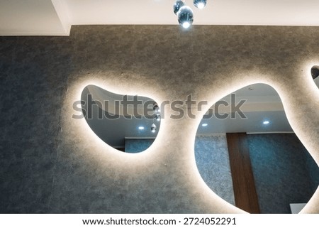 Organic Backlit Mirrors Casting Soft Glow On Textured Gray Wall, Irregular Shapes Form Sculptural Focal Point With LED Halo, Reflected Curtain And Recessed Lights Add Depth, Contemporary Boutique