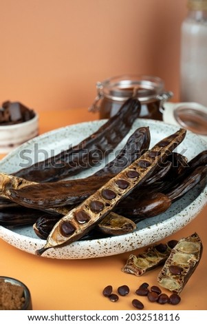 Organic carob pods, seeds in a ceramic plate on a beige background, locust bean healthy food, Ceratonia siliqua harnup. Natural vegan eating. creative food background. Copy space. Top view.