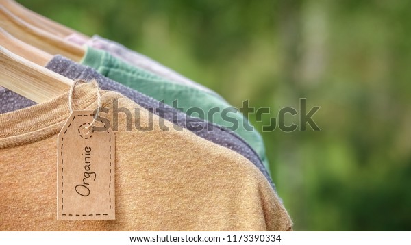 Organic clothes. Natural colored t-shirts\
hanging on wooden hangers in a row. Eco textile tag. Green forest,\
nature in background.