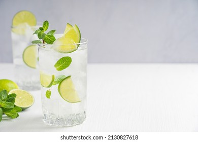 Organic cold refreshing lemonade drink or cocktail made of sparkling water, lime slices and fresh green mint leaves served in tall drinking glass with ice cubes on white wooden table with copy space