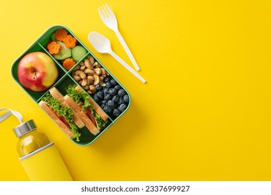 Organic school lunch presentation. Top view exhibiting eco-conscious lunchbox filled with nutritious treats, fruits, veggies, berries, nuts, water bottle, cutlery on yellow surface with room for ad