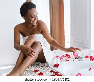 Organic spa, skincare and beauty care, weekend relaxation. Happy young african american lady in white towel puts petals into bathtub in white bathroom interior in daylight, empty space, square