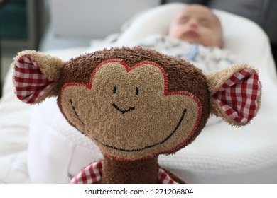 Organic Stuffed Monkey Toy With Blurred Face Baby In The Background Laying On A White Bed