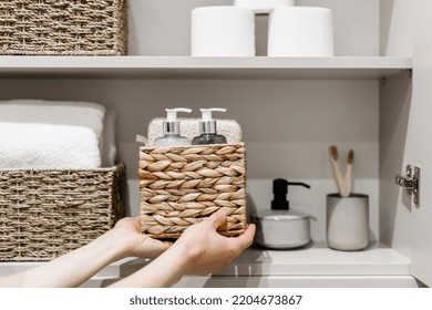 Organization of space in the bathroom cabinet. Cropped view of woman putting wicker box with bath sponge, shampoo, soap dispenser bottle and other cosmetics products in closet