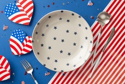 Organize Menu For Fourth Of July Gathering. Top-view Shot Of Festive Table Featuring Empty Plate, Silverware, Heart-shaped Flags, And Vibrant Confetti On USA Flag Inspired Backdrop With Space For Text