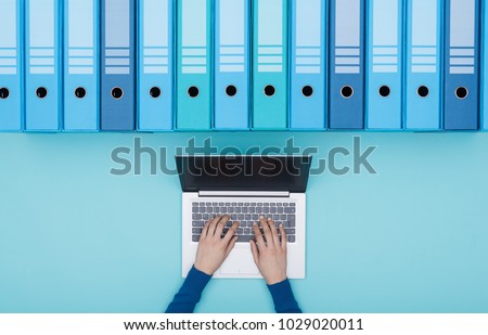 Organized archive with ring binders and woman searching for files in the database using a laptop, top view