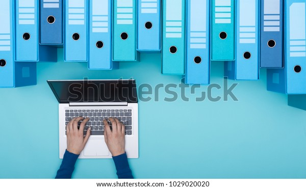 Organized archive with\
ring binders and woman searching for files in the database using a\
laptop, top view