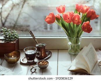 Oriental Coffee Cooked In Traditional Turkish Copper Coffee Pot With Flowers On Window Sill. Wooden Windowsill With Bunch Of Tulips Book.Cozy Scene, Hygge Concept. Cold Rainy Day In Spring.