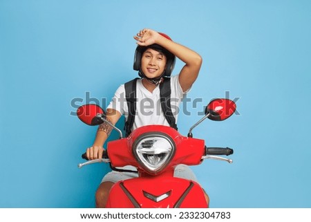 Oriental delivery guy in red helmet sits on red scooter in studio, one hand on handle bar another on his helmet, fast drive concept, copy space