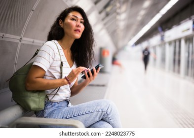 Oriental woman sitting on bench in metro station and using her smartphone while waiting for train. - Shutterstock ID 2248023775