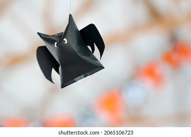 Origami Bat Made Of Black Paper Hanging On A Rope For Halloween Decorations. Dark Paper Ghost Halloween Party Concept Origami Paper Bat. The Figure Of Black Paper Bat Flying Over Room. Space For Text.