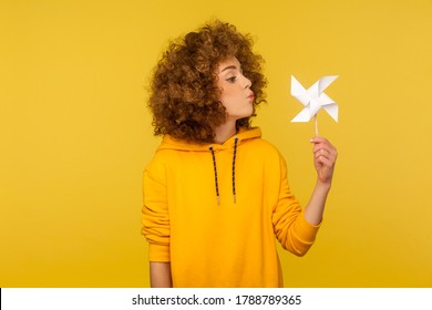 Origami hand mill. Portrait of carefree childish curly-haired woman in urban style hoodie blowing at paper windmill, playing with pinwheel toy on stick. studio shot isolated on yellow background