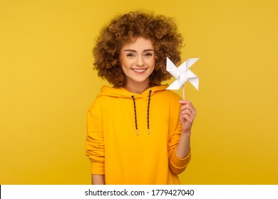 Origami Hand Mill. Portrait Of Happy Curly-haired Woman In Urban Style Hoodie Smiling Carefree And Holding Paper Windmill, Pinwheel Toy On Stick. Indoor Studio Shot Isolated On Yellow Background