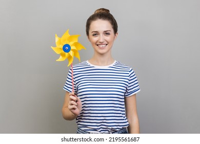 Origami Hand Mill. Portrait Of Happy Woman Wearing Striped T-shirt Smiling Carefree And Holding Paper Windmill, Pinwheel Toy On Stick. Indoor Studio Shot Isolated On Gray Background.