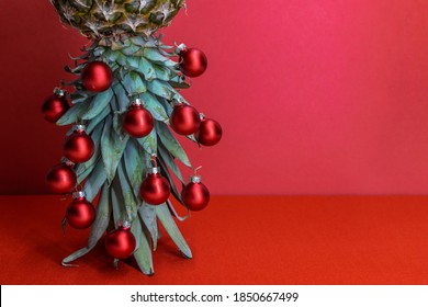 Original Christmas tree concept. Red shiny glass baubles hangs on the leaves of ripe pineapple standing upside down. Red background with copy space for your text.