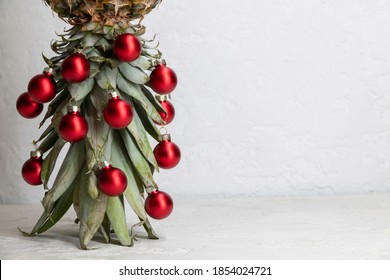 Original Christmas tree concept. Red shiny glass baubles hangs on the leaves of ripe pineapple standing upside down. Gray concrete background with copy space for your text. Selective focus.