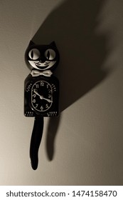 The Original Classic Kit Cat Clock, The Clock Shaped Like A Cat In A Tuxedo, Hangs On A Wall On Left-hand Side Of Frame With Dramatic Lighting. 

