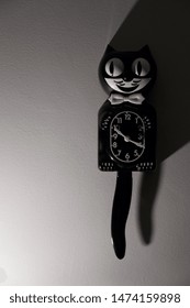 The Original, Classic Kit Cat Clock, The Clock Shaped Like A Cat In A Tuxedo, Hangs On A Wall On Right-hand Side Of Frame With Dramatic Lighting. 
