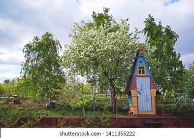 The original colorful wooden garden toilet in the village, on the site against the backdrop of tall tall green leafy trees. Garden exterior. Russian outdoor toilet. 