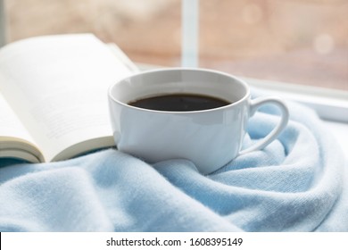 Original photograph of a large cup of coffee surrounded by a soft blue throw with a book in the window sill on a cold day