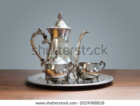 Ornate 4-piece tea set on table. Silver or silver plated tea pot, sugar bowl and cream or milk jug. Ornamental silverware to serve hot tea or coffee. Selective focus.