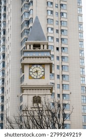 Ornate clock tower with a classical design and pointed spire standing in stark contrast against the backdrop of a modern high-rise residential apartment building, viewed through bare tree branches.