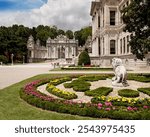 Ornate gardens of Dolmabahçe Palace featuring manicured flower beds, a lion statue, and the grand imperial gate under a partly cloudy sky.
