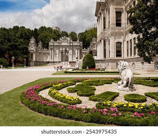 Ornate gardens of Dolmabahçe Palace featuring manicured flower beds, a lion statue, and the grand imperial gate under a partly cloudy sky.
 - Powered by Shutterstock