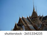 Ornate rooftops and golden spires of the Grand Palace in Bangkok, Thailand, captured under a vibrant blue sky, showcasing traditional Thai architecture.                      