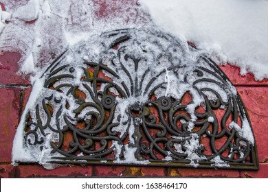 Ornate Rubber Welcome Door Mat In Snow With Ice And Shoe Prints On Grungy Painted Red Brick Outdoor Surface - Top Down View - Closeup