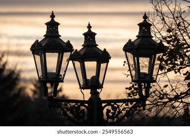 Ornate street lamps silhouetted against a warm sunset sky, Decorative ironwork with detailed patterns, Three ornate, vintage-style street lamps - Powered by Shutterstock