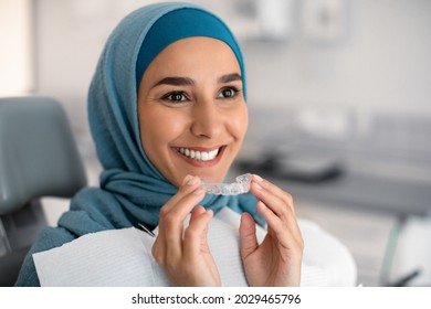 Orthodontics Concept. Smiling Muslim Woman In Hijab Holding Invisalign Or Invisible Braces While Sitting At Dentists Chair In Clinic, Islamic Female Using Clear Dental Aligner For Teeth Correction