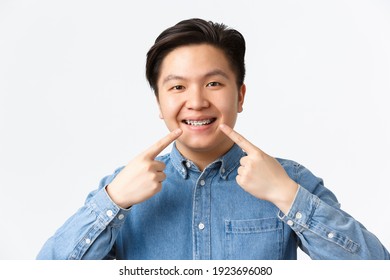 Orthodontics And Stomatology Concept. Close-up Shot Of Happy Smiling Asian Man Recommend Dental Clinic, Pointing Fingers At Teeth Braces With Satisfied Expression, White Background