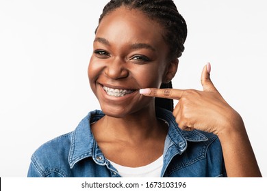 Orthodontics Treatment. Cheerful African Girl In Dental Braces Pointing Finger At Smile And Teeth With Brackets Over White Studio Background.