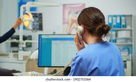 Orthodontist Nurse Talking At Phone Making Dental Appointments In Modern Equipped Office, While Specialist Dentist With Face Mask Is Examining Patient With Toothache Sitting On Stomatological Chair.