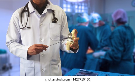 Orthopedic doctor showing model of Total knee replacement with blurred background of surgical team performing surgery in operating room. Medical technology concept. 