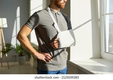 Orthopedic Emergency. Injured Man Who During Rehabilitation At Home Wearing Medical Support Brace And Sling Bandage Immobilizer On His Arm. Young Man With Broken Arm Looks Thoughtfully Out Window.