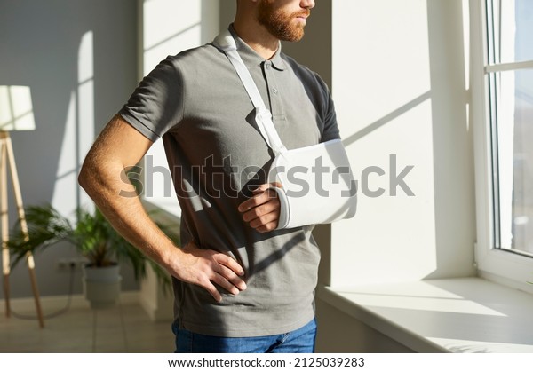 Orthopedic emergency. Injured man who during\
rehabilitation at home wearing medical support brace and sling\
bandage immobilizer on his arm. Young man with broken arm looks\
thoughtfully out\
window.