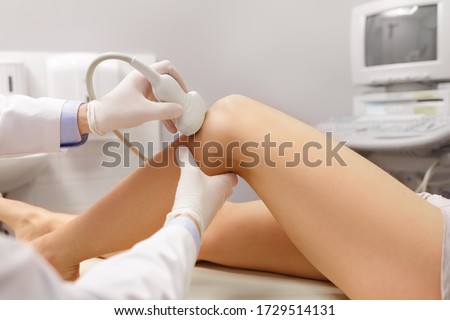 Orthopedist doctor doing ultrasound examination of patient's knee in his office. Young woman passing ultrasound scan in clinic. Doctors work. Medical research.