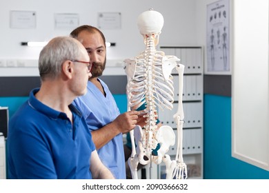 Orthopedist explaining spinal cord on human skeleton to retired patient in physiotherapy office. Chiropractor doing presentation of spine bones and anatomy to old man for osteopathy - Powered by Shutterstock