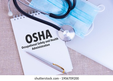 OSHA Occupational Safety And Health Administration, The Text Is Written In A Notebook, Next To A Pen, A Disposable Medical Mask And A Laptop On A Linen Background.