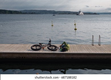 Oslo, Norway - 20 May 2017 - Man Lying Down Beside His Fat Bike And Checking His Phone On Wood Jetty In The Oslo Fjord On Sunny Summer Day