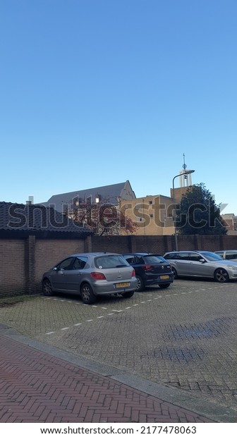 Oss,\
The Netherlands - December 24 2020: Some cars parked in a small\
parking lot belonging to the big church of Oss, The Netherlands. A\
building from a building company in the\
background.