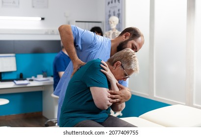 Osteopathic Assistant Cracking Back And Shoulder Bones For Senior Woman At Chiropractic Consultation. Chiropractor Giving Assistance To Elder Patient For Recovery And Rehabilitation