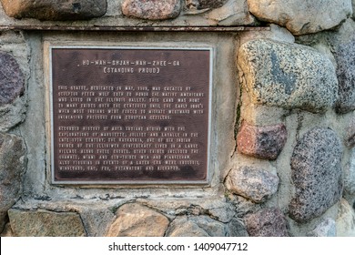 Ottawa, IL / USA - May 25, 2019: A Plaque At The Base Of A Large Wooden Statue Of Native American HO-MAH-SHJAH-NAH-ZHEE-GA, Standing Proud, By Peter Wolf Toth On The Banks Of The Illinois River.