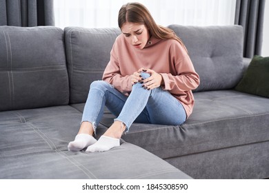 Ouch! Young Caucasian Female Touching Her Sore Knee With A Face Distorted In Pain While Sitting At Thesofa At Home. Health And Illness Concept