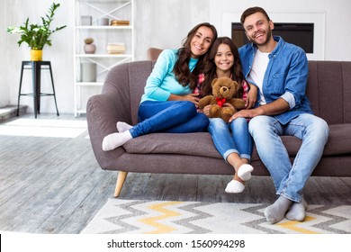 Our Family Portrait. Full-length Family Picture Of Mom, Dad And Their Cute Daughter With A Teddy Bear, Smiling And Hugging On The Couch In The Cozy Home Atmosphere.